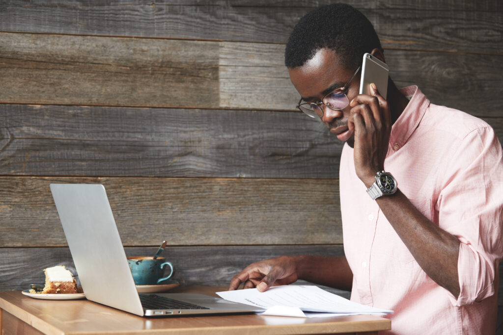 Young African executive officer in formal shirt examining a financial report, making phone calls to
